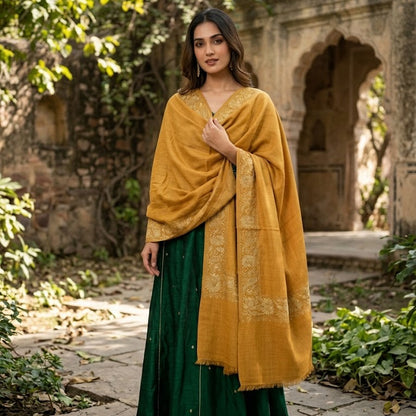 A woman stands outdoors in a stone courtyard wearing a dark green skirt, a mustard yellow shawl draped over her shoulders, and beige shoes. Lush green foliage and an old stone archway are visible in the background.