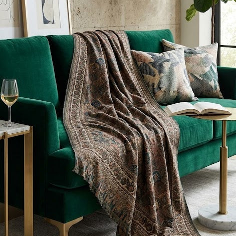 A dark green velvet sofa in a styled living room setting, draped with a brown patterned throw blanket. The scene includes decorative pillows, a side table with a wine glass, framed wall art, a potted plant, and a light-colored stone wall background.