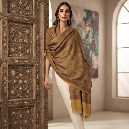 A woman models a draped mustard yellow and brown shawl with subtle paisley patterns, paired with white trousers and beige heels, standing next to an ornate carved wooden door in a warm, elegant room.