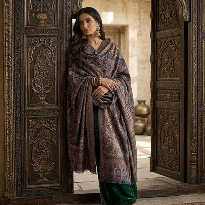 A woman stands in an ornate carved wooden doorway wearing a dark patterned shawl draped over a green skirt. The setting features intricate wood carvings and a traditional patterned rug.