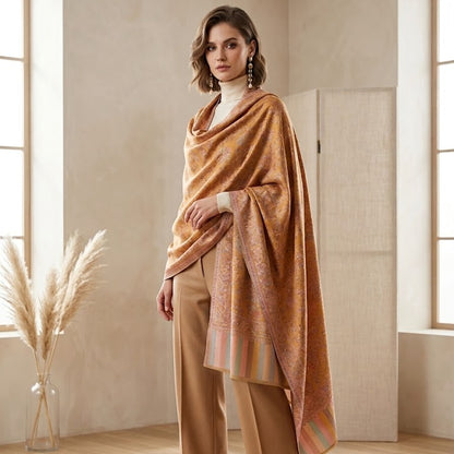 A woman models a luxurious draped shawl in a warm camel-brown and gold tone with subtle striped accents, styled over a white mock neck and tan trousers in a bright, modern room.