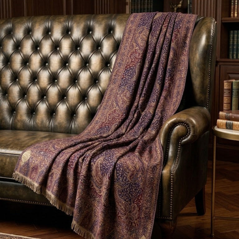 A patterned throw blanket draped over a tufted brown leather sofa in a warm, wood-paneled room with bookshelves and a rug.