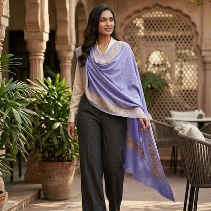 A woman walks through an outdoor courtyard with sandstone arches and plants, wearing a lavender draped shawl with gold border detailing over a cream top and dark gray wide-leg trousers.