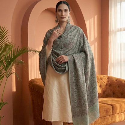 A woman models a light sage green shawl with a subtle paisley pattern, draped over a cream-colored kurta and trousers set, standing in a warm-toned room with terracotta arches and a tufted sofa.