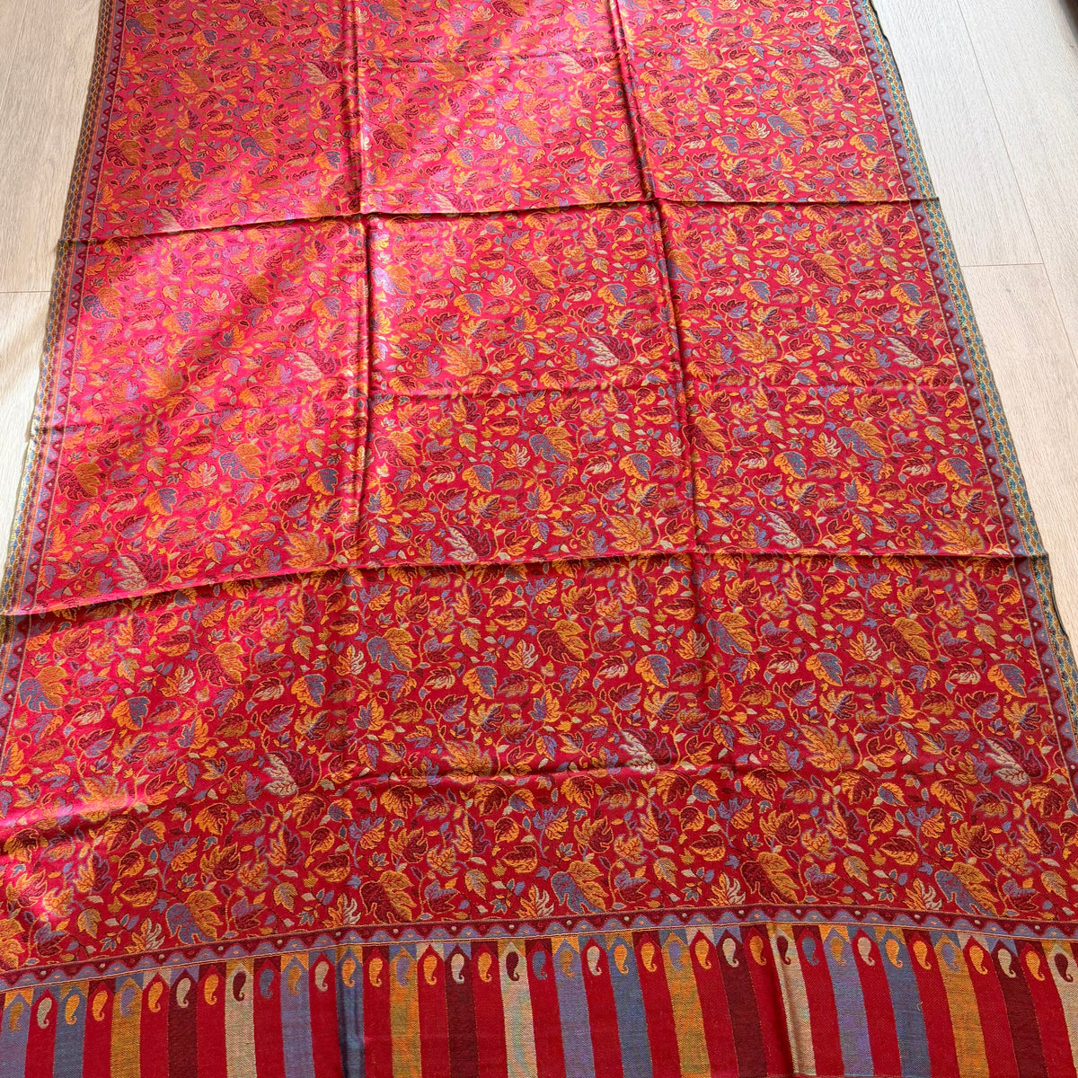 A long red patterned textile, likely a shawl or scarf, laid flat on a light wood floor. The fabric features an intricate floral and paisley pattern in red, orange, yellow, and blue, with a striped border in red, blue, and gold at the bottom.