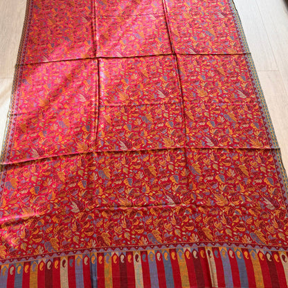 A long red patterned textile, likely a shawl or scarf, laid flat on a light wood floor. The fabric features an intricate floral and paisley pattern in red, orange, yellow, and blue, with a striped border in red, blue, and gold at the bottom.