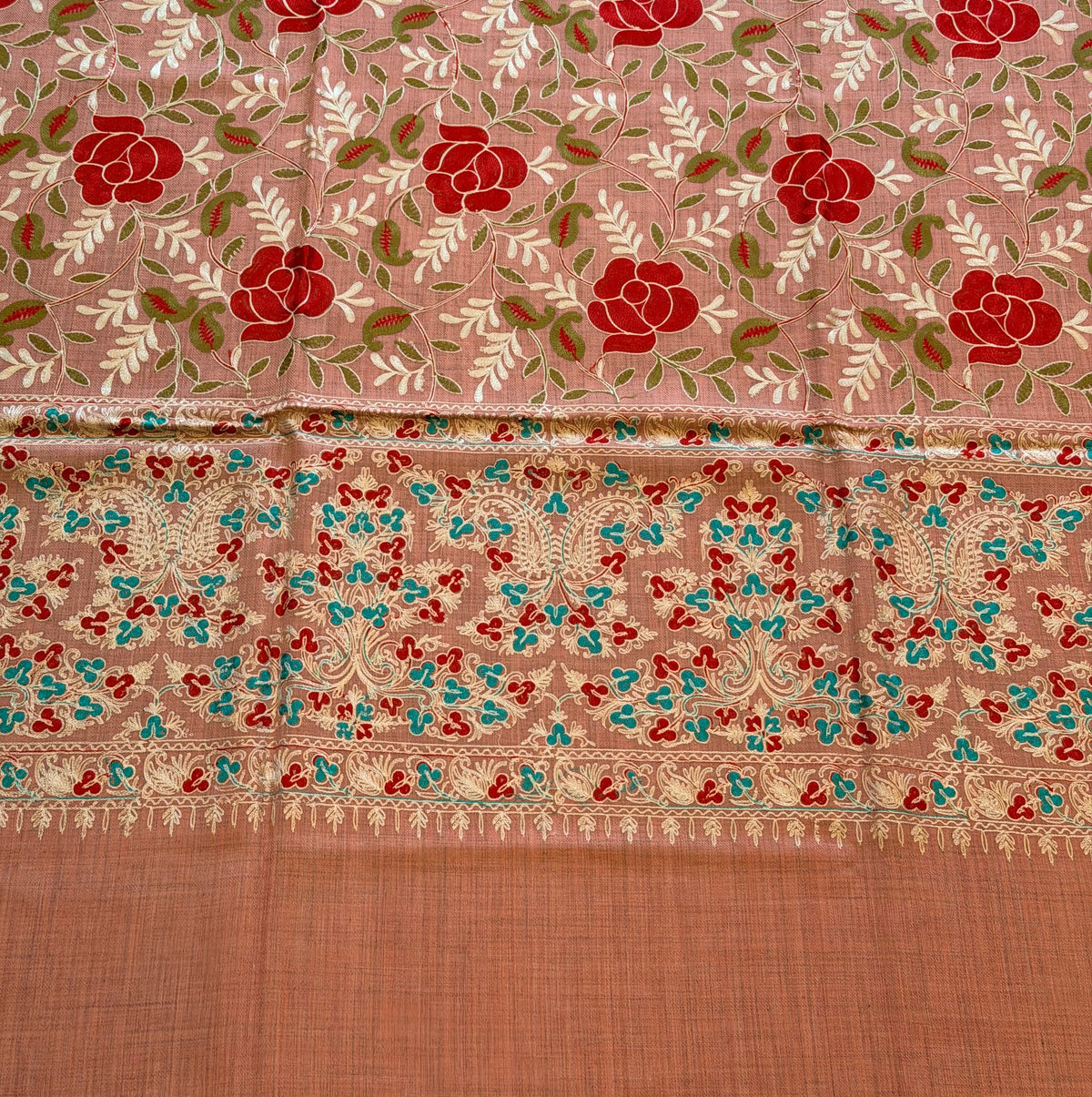 A close-up of a patterned fabric, likely a saree, featuring a dusty pink body with red rose motifs, a detailed multicolor border, and a solid peach-colored pallu.