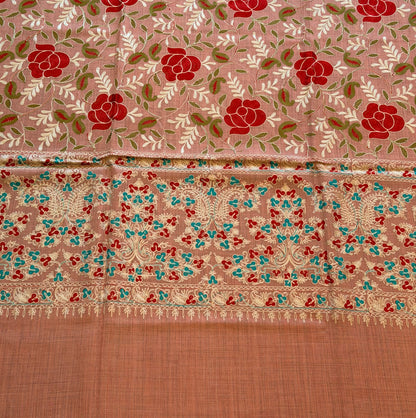 A close-up of a patterned fabric, likely a saree, featuring a dusty pink body with red rose motifs, a detailed multicolor border, and a solid peach-colored pallu.