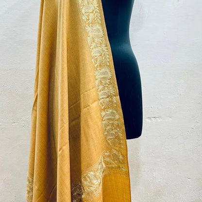A mustard yellow shawl with an intricate beige floral border, draped over a black mannequin torso against a textured off-white wall.