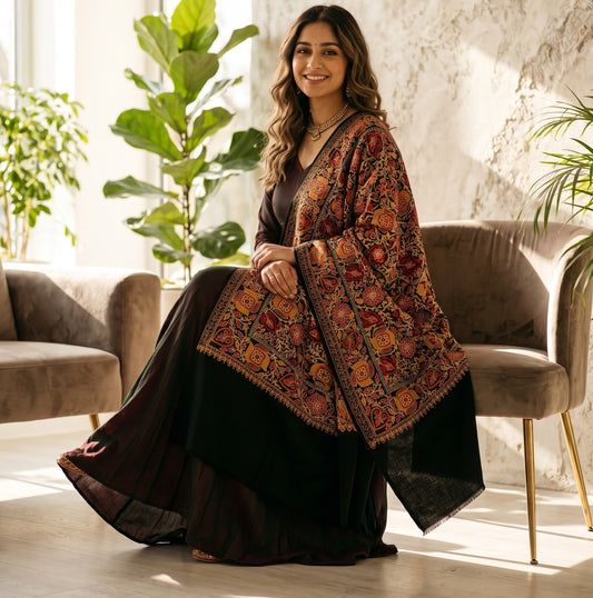 A woman sits on a plush armchair in a bright, modern room, wearing a black skirt and a dark brown shawl with intricate red, orange, and gold floral patterns.