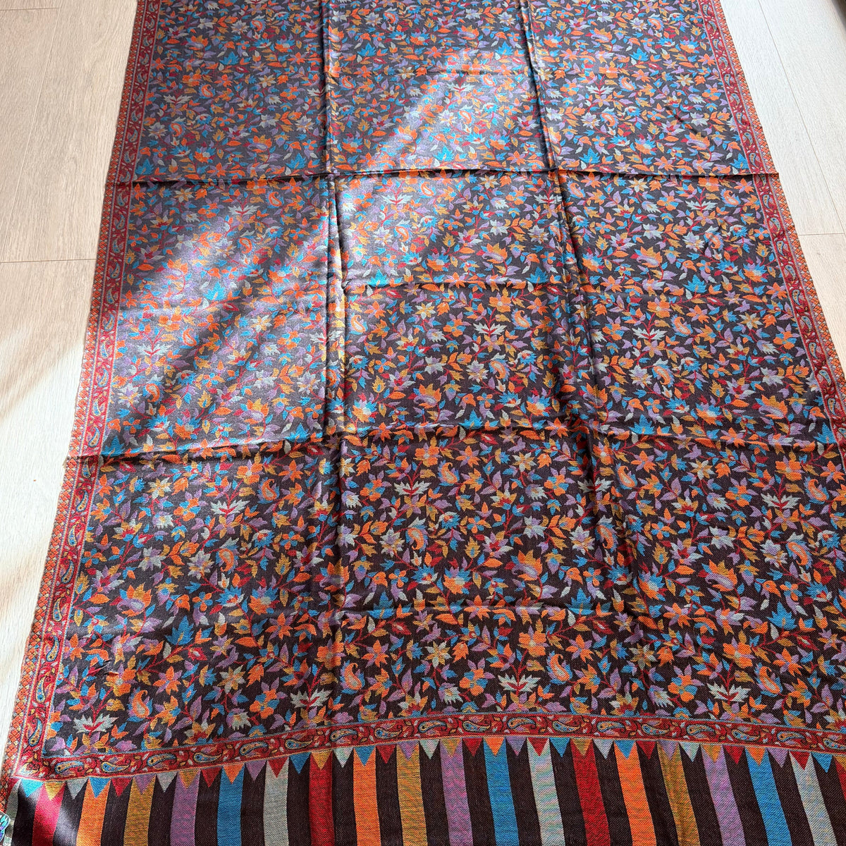 A large rectangular patterned textile laid flat on a light wood floor, featuring a dense floral print in blue, orange, red, and yellow on a dark background, with striped multicolor borders and fringe along the bottom edge.