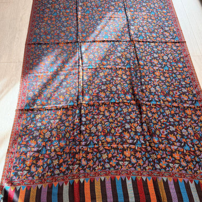 A large rectangular patterned textile laid flat on a light wood floor, featuring a dense floral print in blue, orange, red, and yellow on a dark background, with striped multicolor borders and fringe along the bottom edge.