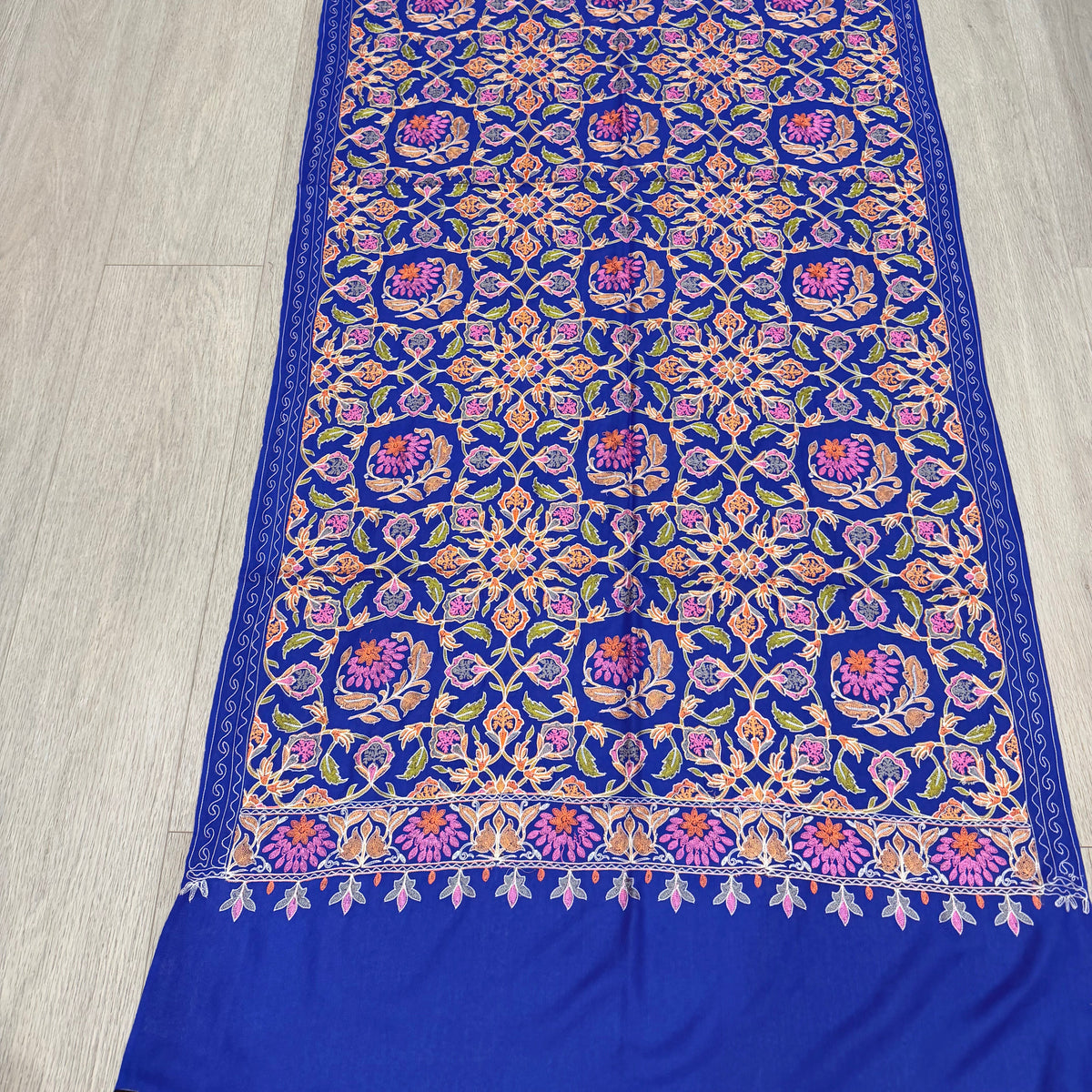 A royal blue fabric piece, likely a sari or scarf, featuring an intricate multicolor embroidered pattern with purple, pink, orange, and green motifs, laid flat on a light wood floor.