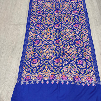A royal blue fabric piece, likely a sari or scarf, featuring an intricate multicolor embroidered pattern with purple, pink, orange, and green motifs, laid flat on a light wood floor.