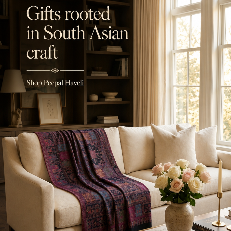 A styled living room scene with a cream upholstered sofa, a dark patterned throw blanket, a wooden coffee table with flowers and books, and large windows with curtains. Text on the left reads 'Gifts rooted in South Asian craft' and 'Shop Peepal Haveli'.