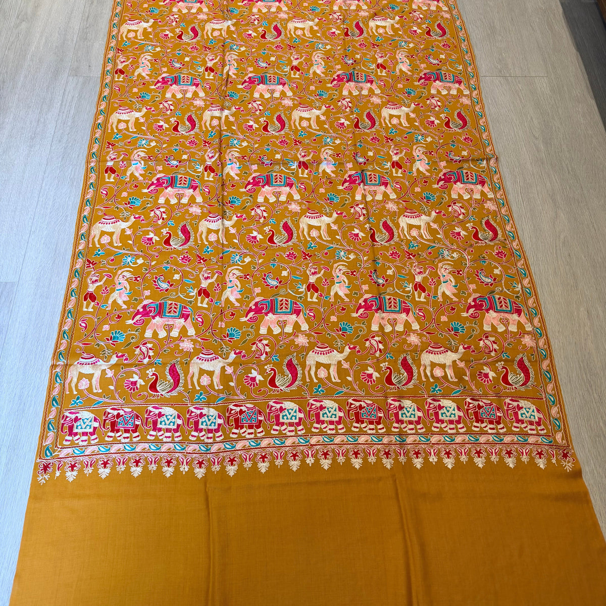A mustard yellow Banarasi silk saree with intricate multicolor embroidery depicting elephants, lions, and floral motifs, laid flat on a light wood floor.