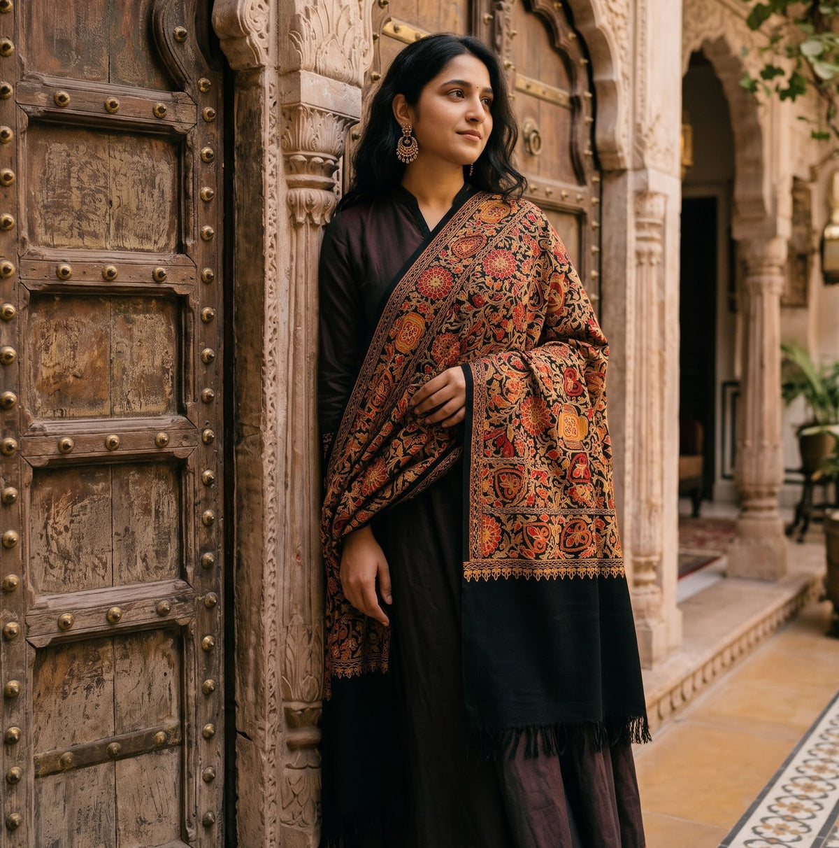 A woman stands in a traditional courtyard wearing a dark brown kurta and a richly patterned orange, red, and black embroidered dupatta with a black border.