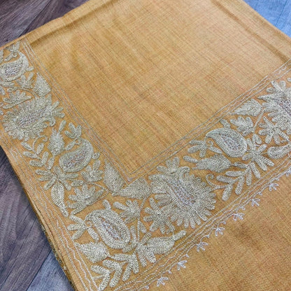 A mustard yellow woven textile, likely a dupatta or scarf, featuring an ornate beige and gold floral paisley border, laid flat on a dark wood floor.