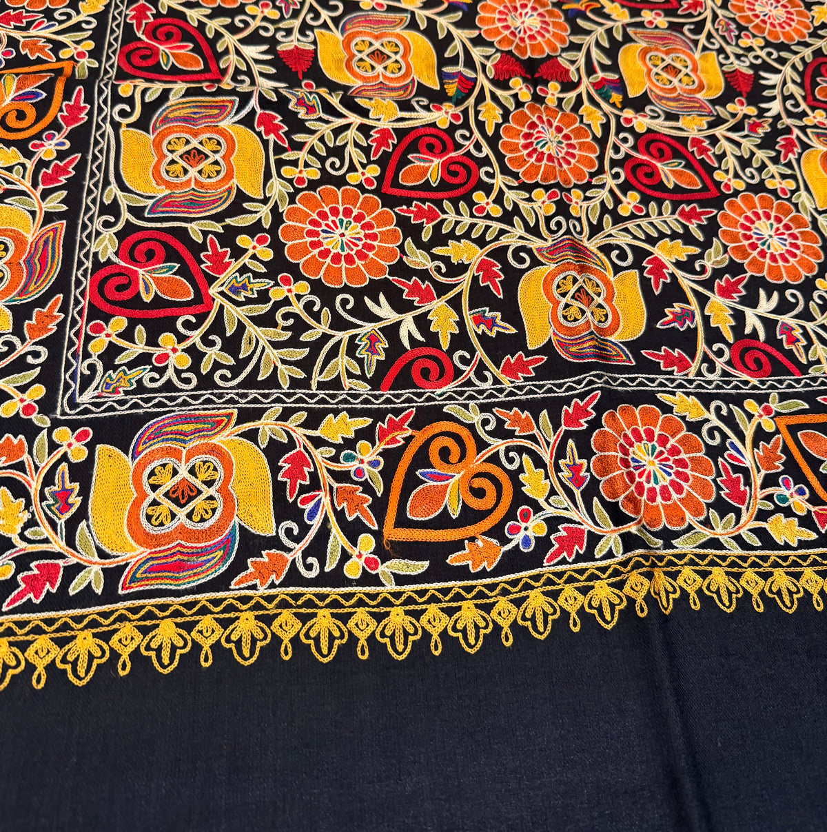 A close-up of a black fabric with dense, colorful hand-embroidered floral and geometric patterns in orange, yellow, red, and green, featuring a decorative border.