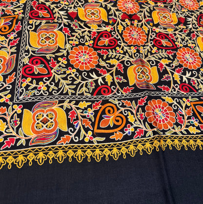 A close-up of a black fabric with dense, colorful hand-embroidered floral and geometric patterns in orange, yellow, red, and green, featuring a decorative border.