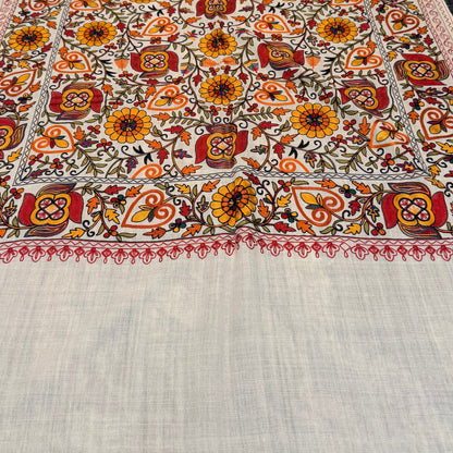 A large rectangular woven textile laid flat on a dark countertop, featuring an ornate multicolor floral and geometric pattern with a wide plain off-white lower section and fringe along one edge.