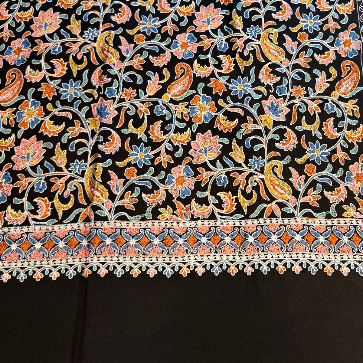 A black fabric textile with an intricate multicolor floral and paisley pattern, laid out on a light wood floor.