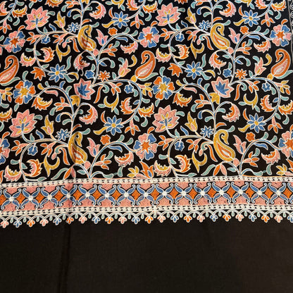 A black fabric textile with an intricate multicolor floral and paisley pattern, laid out on a light wood floor.