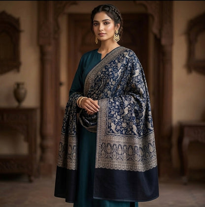 A woman stands indoors wearing a dark teal traditional outfit with a navy blue shawl featuring intricate beige and gold floral patterns and borders. The background shows a softly blurred warm-toned interior with wooden architectural details.