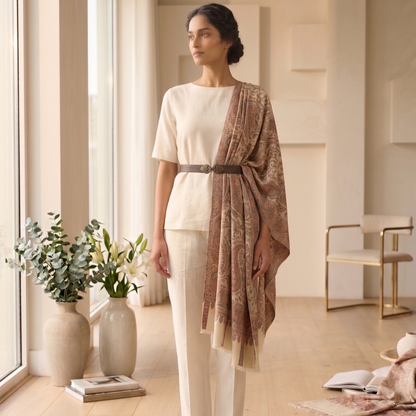 Woman in a white outfit with a brown shawl standing in a bright room with large windows.