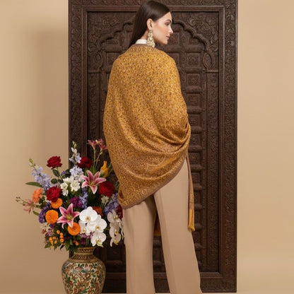 Woman wearing a mustard shawl standing in front of a decorative wooden door with a vase of flowers on a beige background