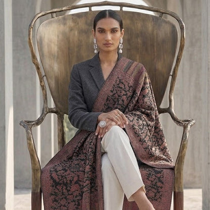 A woman sits in a rustic wooden chair wearing a dark gray blazer, white trousers, and a dark brown patterned shawl draped over her lap, set against a stone archway background.