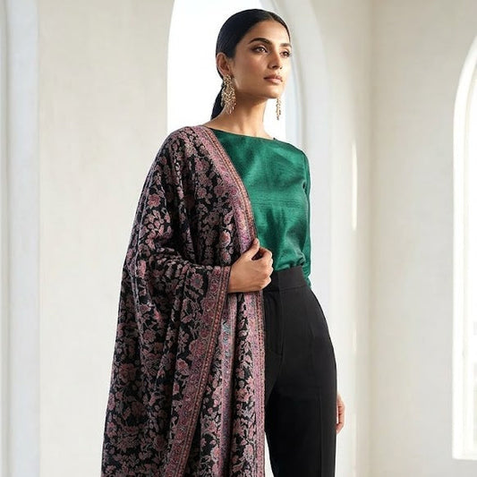 A woman models a traditional South Asian outfit featuring a dark patterned shawl draped over a green top and black trousers, standing in a bright, minimalist white room with arched windows.