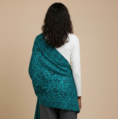 Model wearing a blue patterned faux Pashmina stole over a white top and gray pants on a beige background
