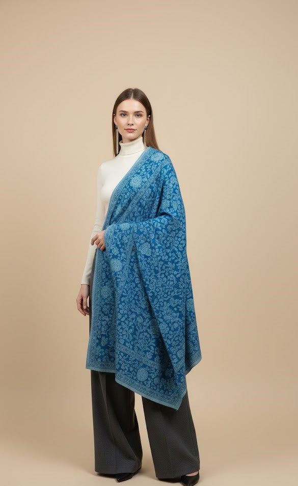 Woman wearing a blue patterned shawl over a white top and black pants on a beige background