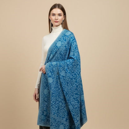 Woman wearing a blue patterned shawl over a white top and black pants on a beige background